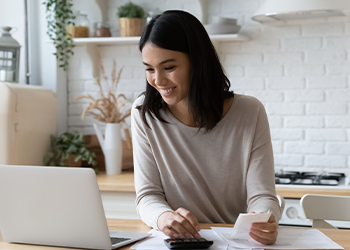 A woman looking at her laptop and smiling. She is holding a pen over a piece of paper.