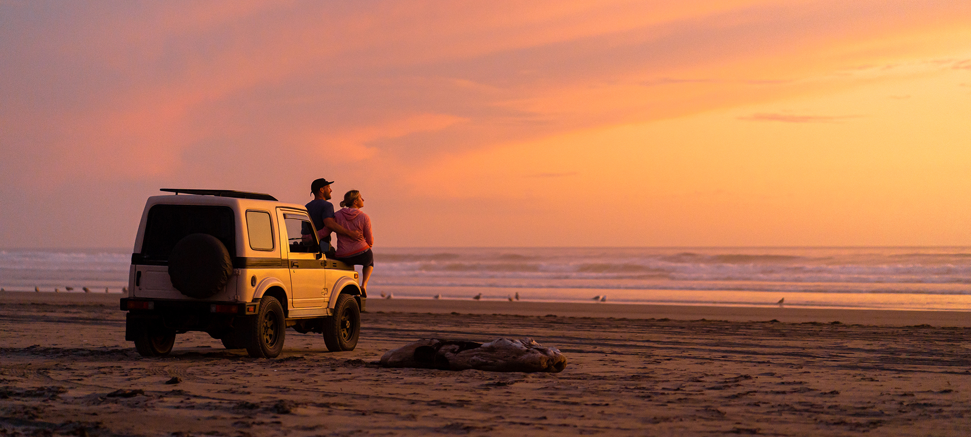 Couple watching sunset from hood of their jeep on the beach